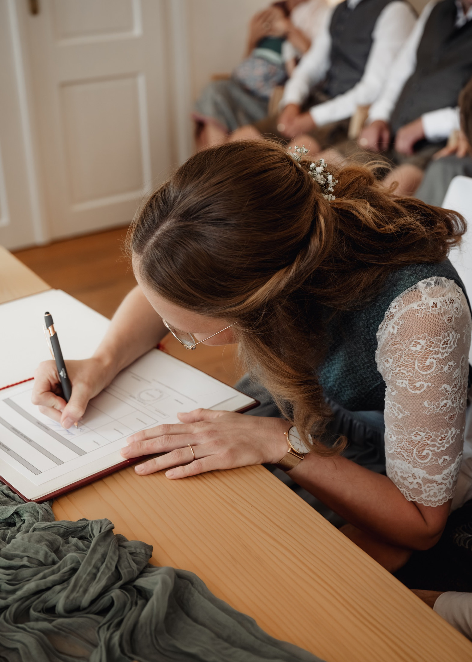 Braut im Dirndl unterschreibt die Heiratsurkunde, Gäste im Hintergrund