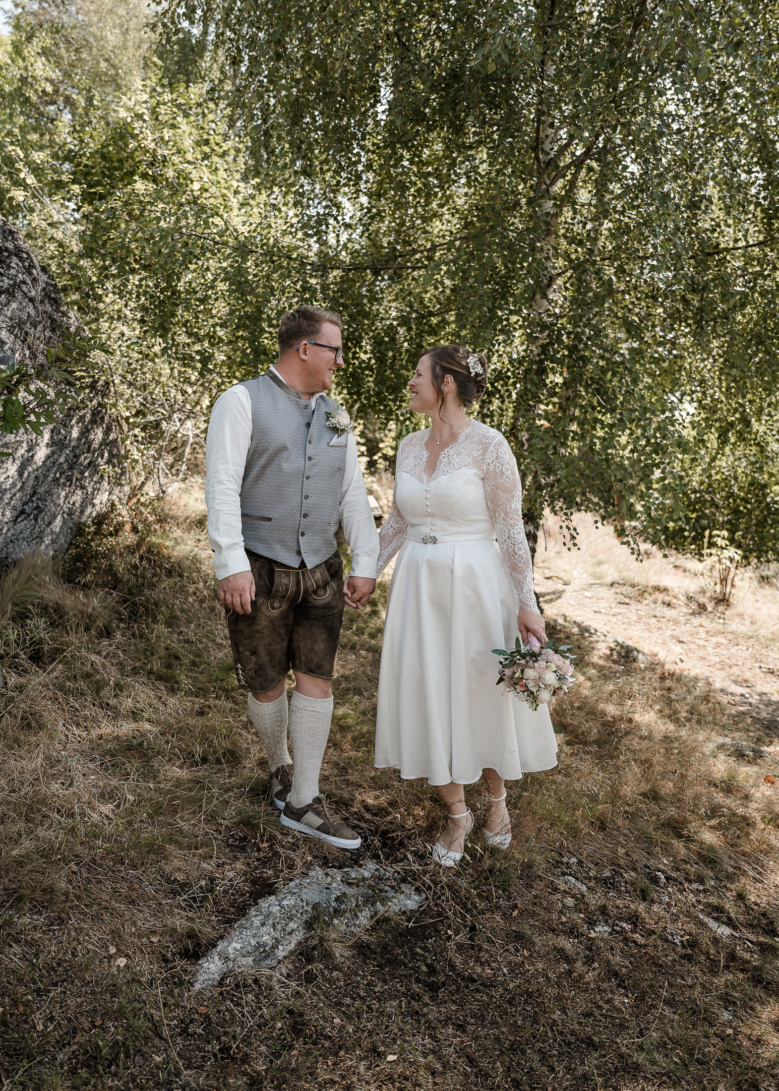 Braut und Bräutigam in Tracht lächeln sich zwischen Birken an