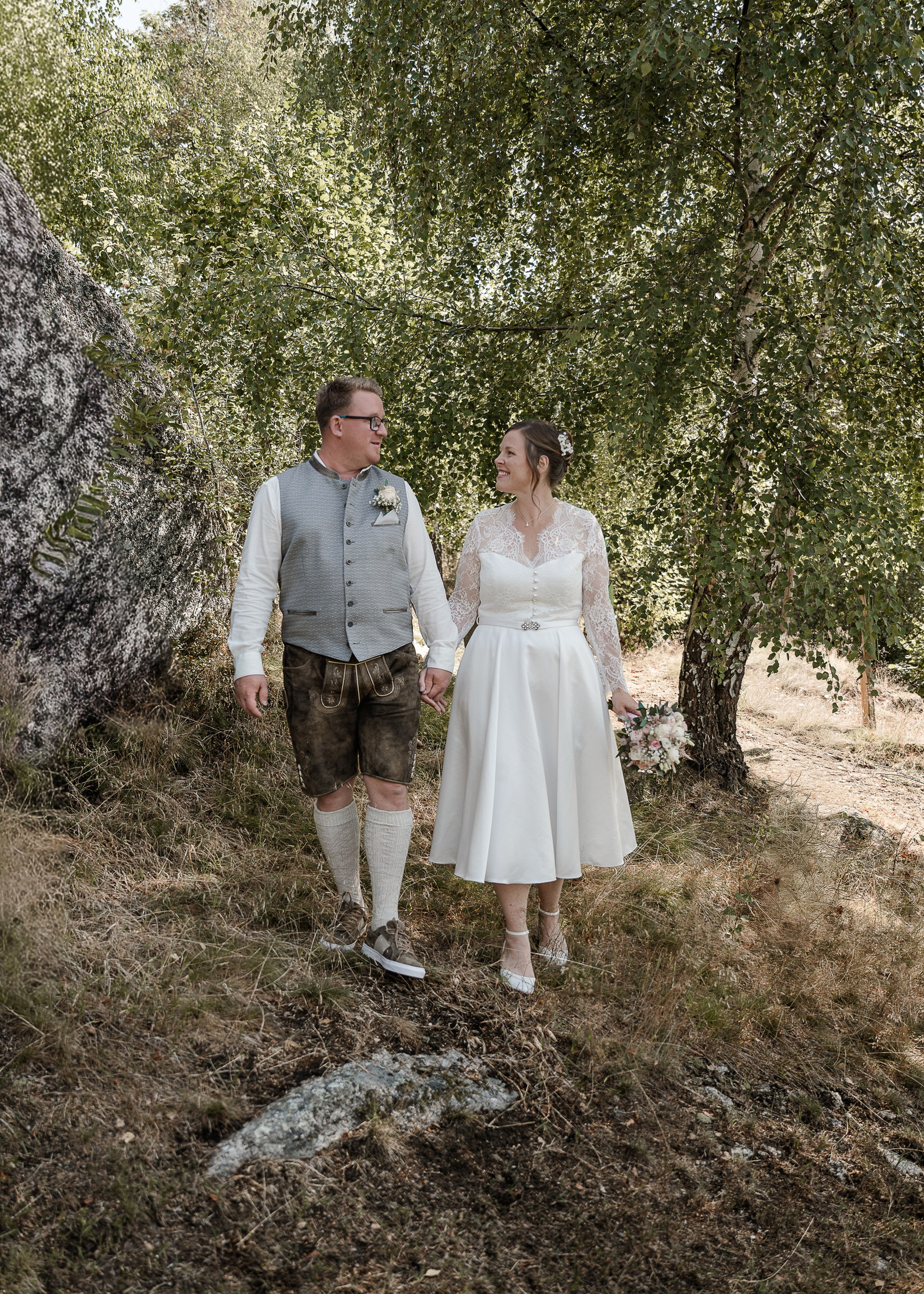 Brautpaar in Tracht geht Hand in Hand zwischen Birken und Fels