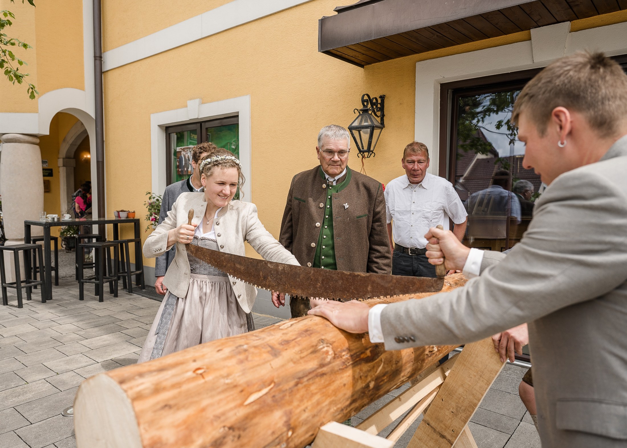 Braut und Bräutigam sägen gemeinsam einen Baumstamm, Gäste schauen zu