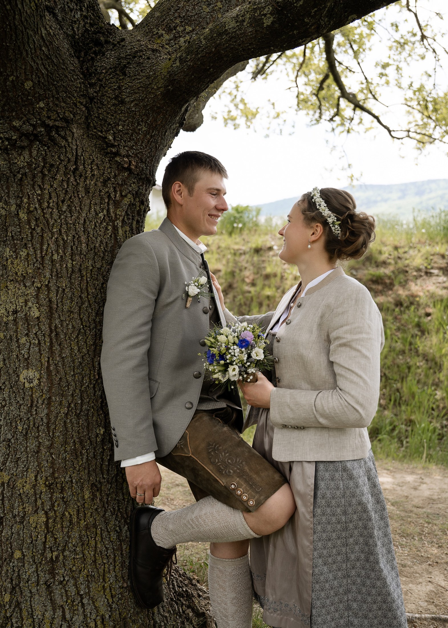 Brautpaar lehnt am Baumstamm und blickt sich verliebt an